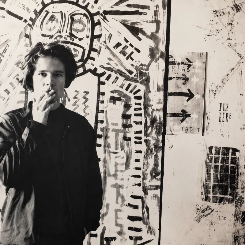L’artiste jeune, cigarette à la main, devant de grands formats peints — noir et blanc