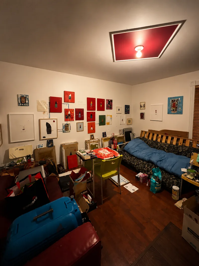 Vue d’ensemble de l’atelier : murs couverts d’œuvres, lit, valises et plafonnier rouge